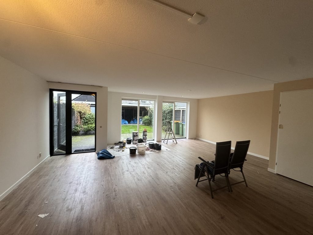 Lege woonkamer met lichte wanden, een houten vloer en verfemmers bij de tuindeuren